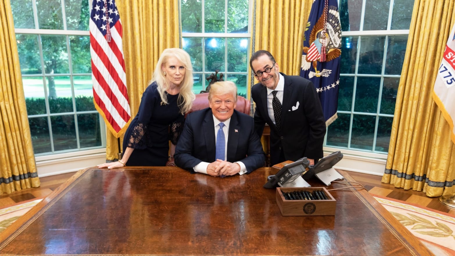 President Donald Trump in the Oval Office, flanked on his left by a man in glasses and his right by a blonde woman.