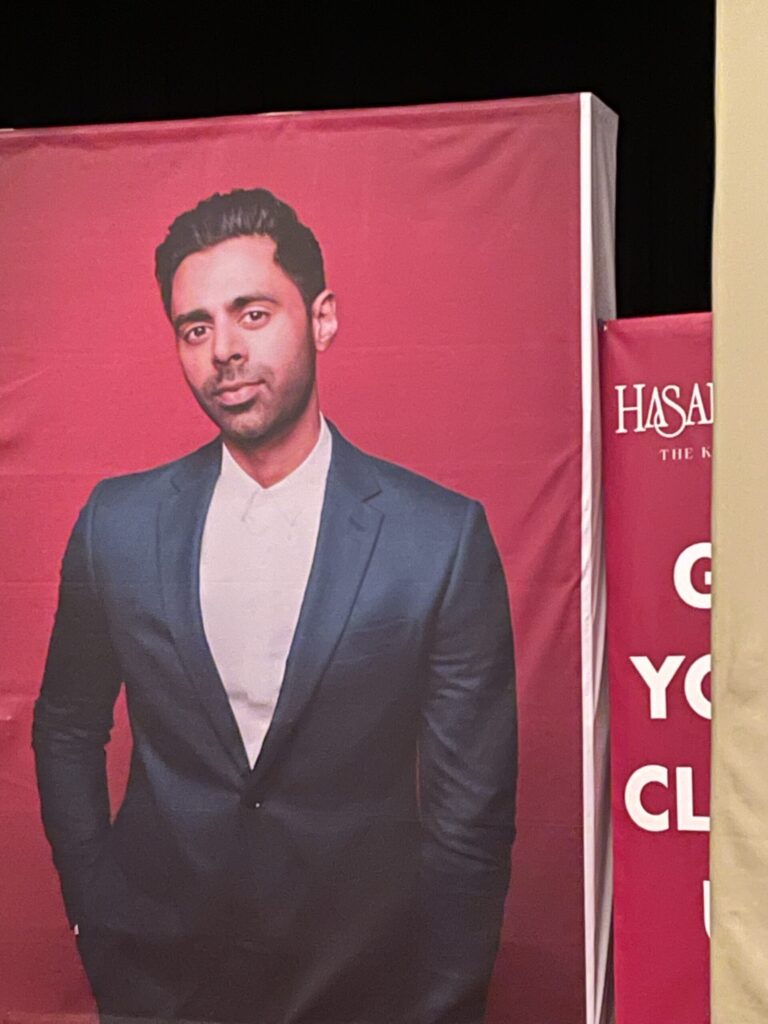 A poster of Hasan Minhaj shows the comedian wearing a dark suit against a bright red backdrop.