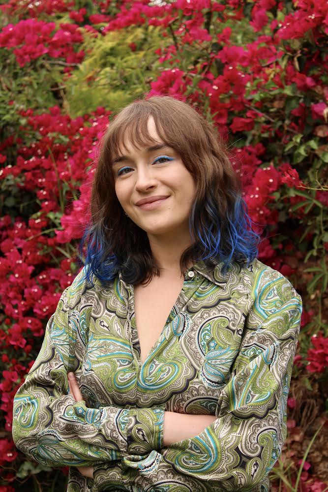 Woman in green dress and blue hair posing in front of red flowers