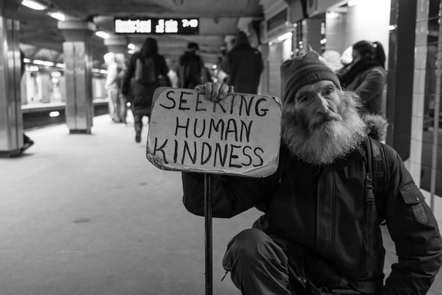 A homeless person holds a sign saying "Seeking Human Kindness" in a subway station
