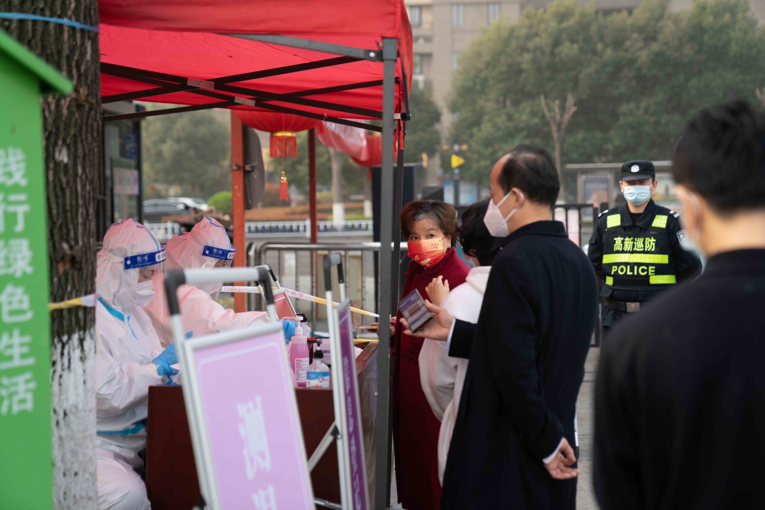 Masked residents stand in line before PPE-clad health workers and police officers, presenting phones to be checked.