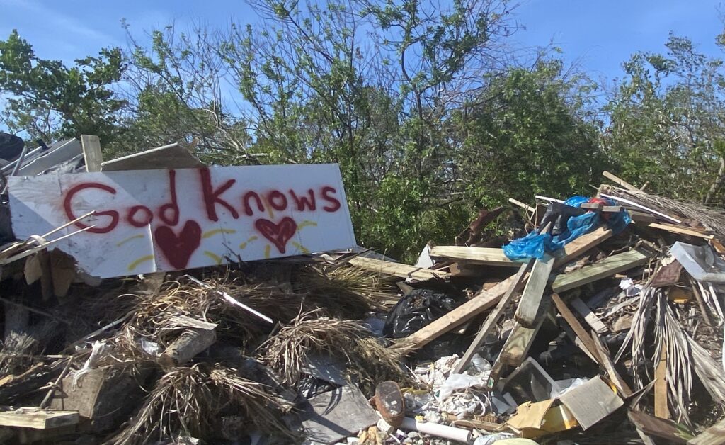 A sign that says "God Knows" sits atop a pile of debris before a row of wild bushes on a sunny day.