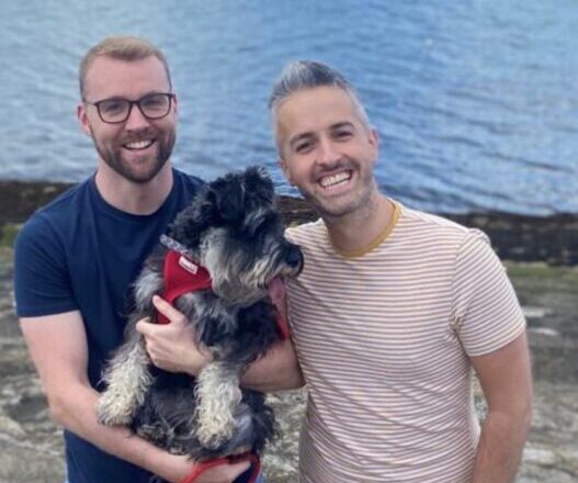 Two happy men posing for a picture with a scruffy schnauzer.