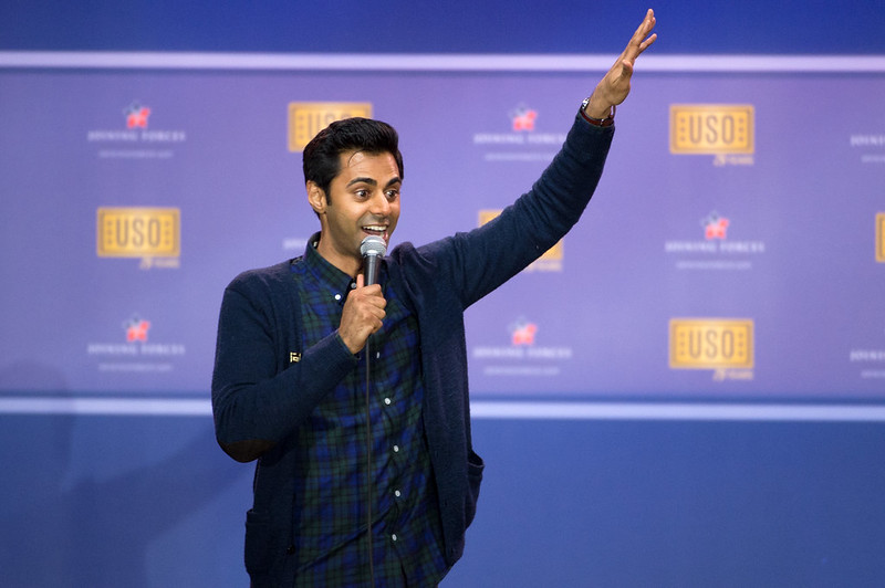 Comedian Hasan Minhaj stands in front of purple backdrop, holding a mic.