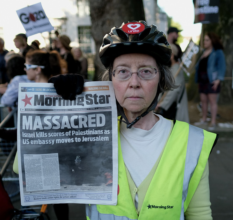 Women in bike helmet and green construction jacket holds up a Morning Star newspaper.