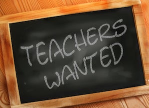 A classroom chalkboard with "teachers wanted" written in white chalk.