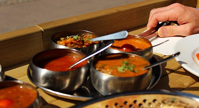 Four Indian curries are placed in steel bowls. One hand is reaching into one of the curries with a spoon.