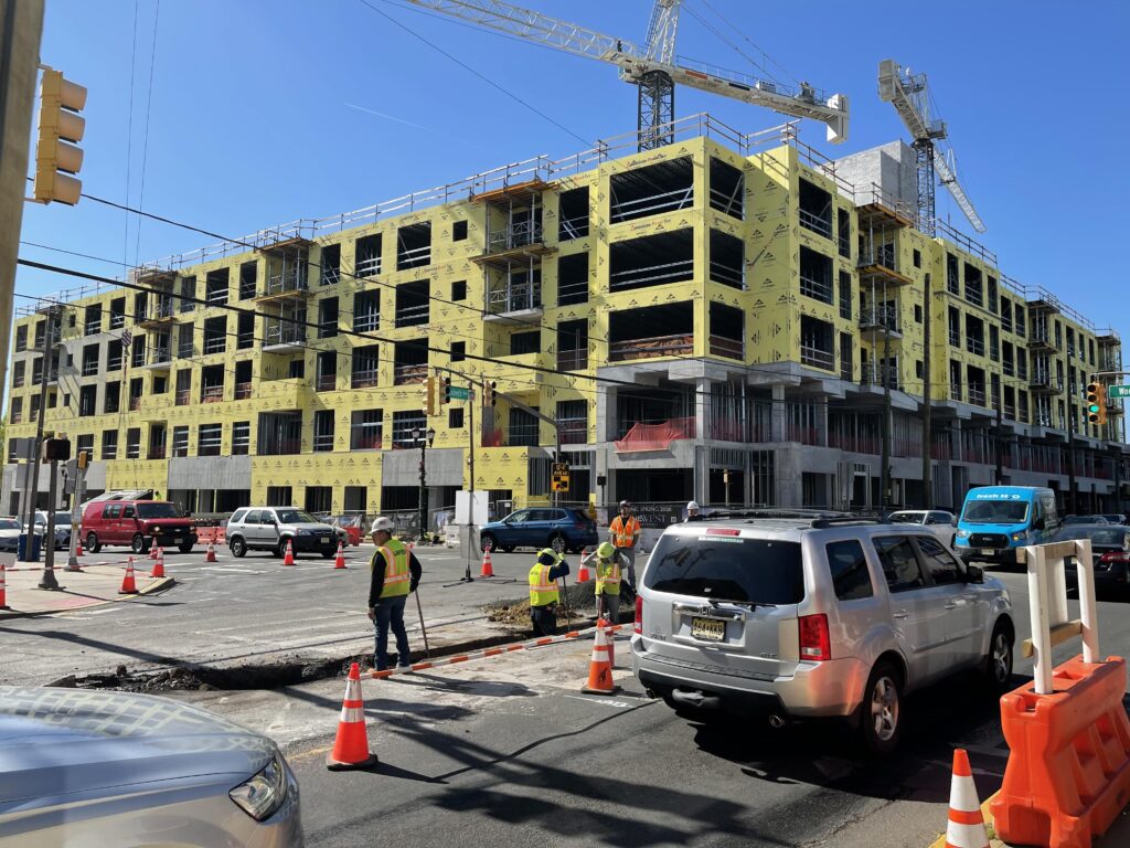 Construction site of the OneWest development in Linden, New Jersey, with workers, traffic cones, and cranes surrounding a multi-story building wrapped in yellow sheathing