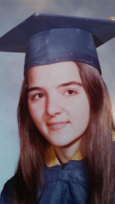 This is a photo of a teenaged girl with long brown hair in dark blue graduation cap and gown. The style and tint of the photo are like what you'd expect to see in a yearbook from the 1970s.