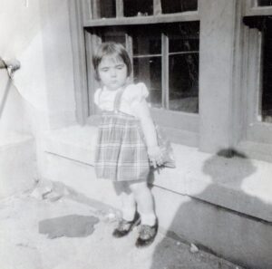 A young girl in a short dress and old-fashioned shoes stands awkwardly perched against a low window sill in what appears to be an old black and white photo.
