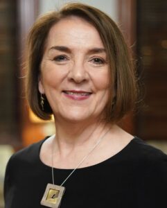 This is a professional business-style headshot of a mature woman with chestnut brown hair, conservative makeup, a big square necklace, and a black shirt. Her haircut is straight and short, somewhat reminiscent of the youthful style she had when she was 3.