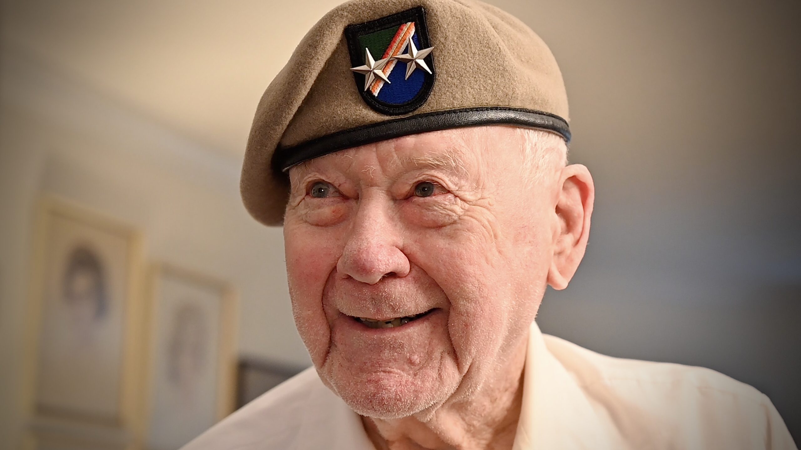 Elderly man (John Raaen) smiling and wearing a tan beret with insignia denoting his final U.S. Army rank of Major General. He retired from the Army in 1979.
