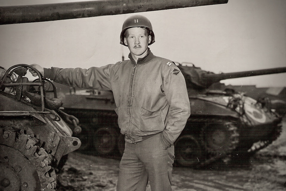 U.S. Army Captain John Raaen wearing a helmet and field jacket stands in front of tanks, resting one arm on a tank, circa 1944.