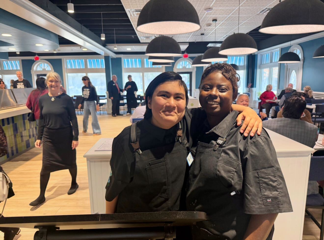 Hugs Cafe Staff Natalie and Kayun smile by hostess stand with Founder Ruth in the background.