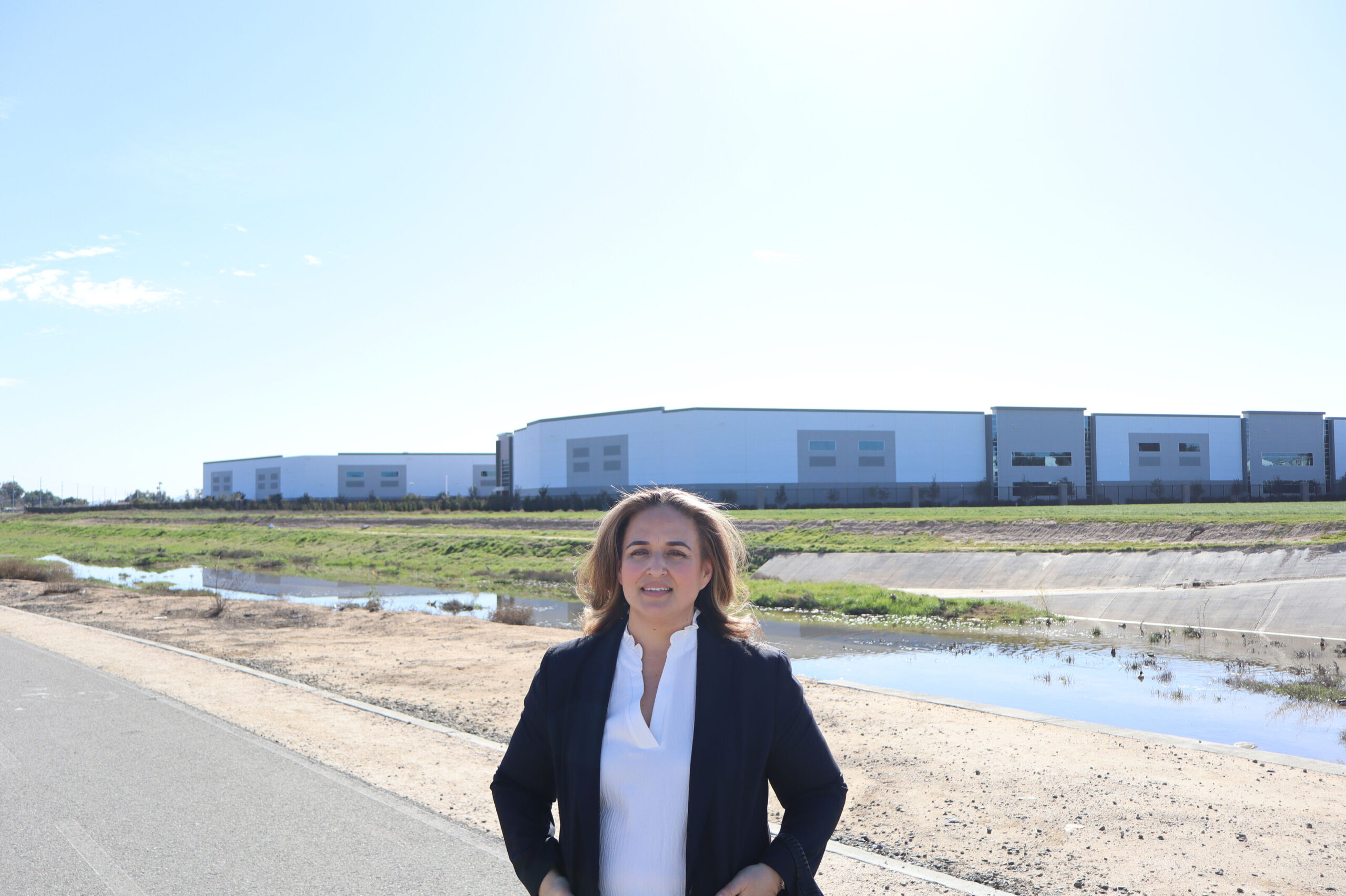 A person in professional attire standing on a road with large warehouse buildings in the background.