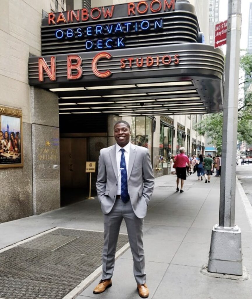 Man in suit stands in front of NBC building sign