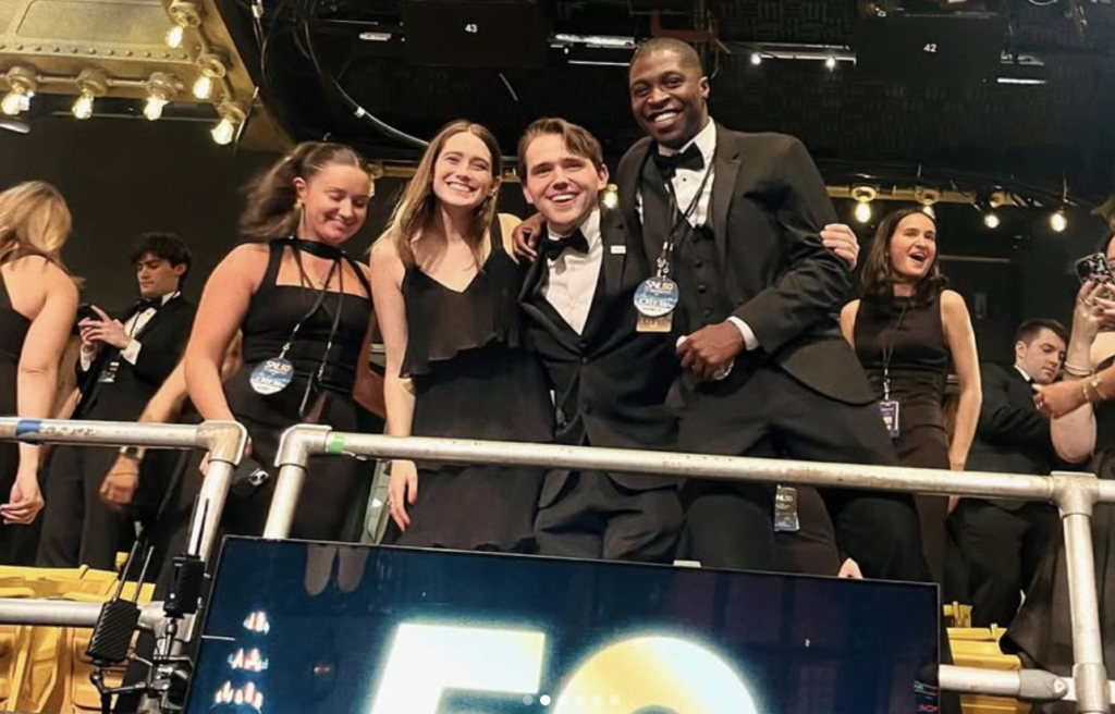 Four workers dressed in formal gear smile at camera in SNL studio